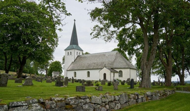 Vit medeltida kyrka med spira och kyrkogård bakom en stenmur